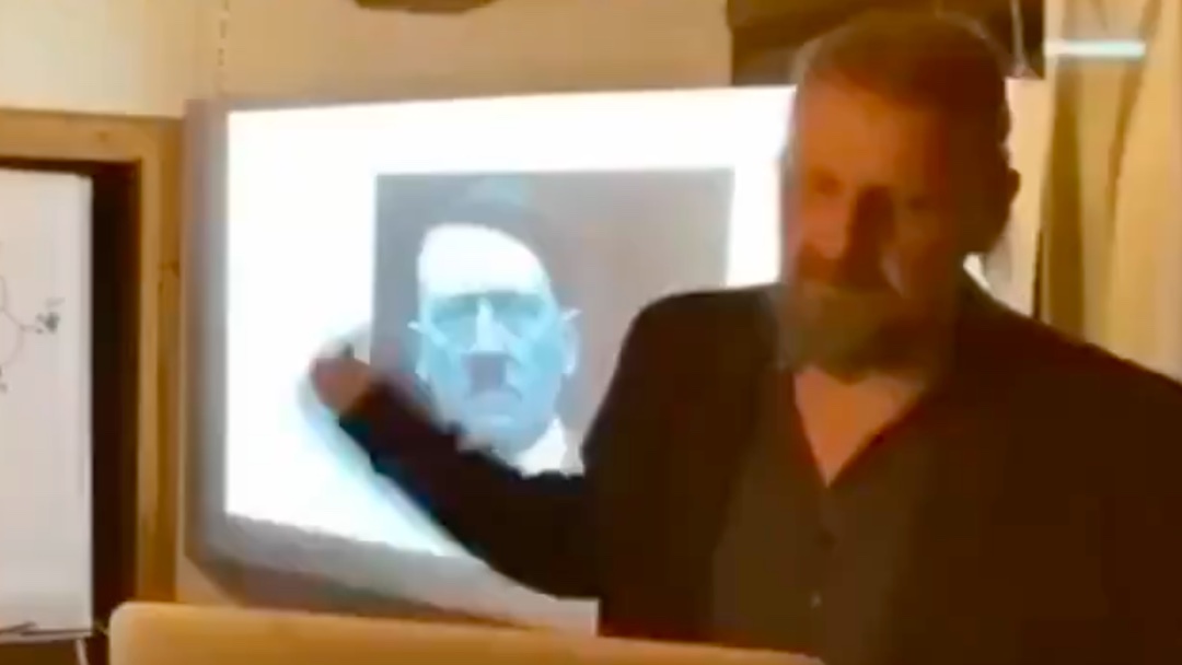 7. Dr. Noack points to an image of Adolf Hitler on the screen.